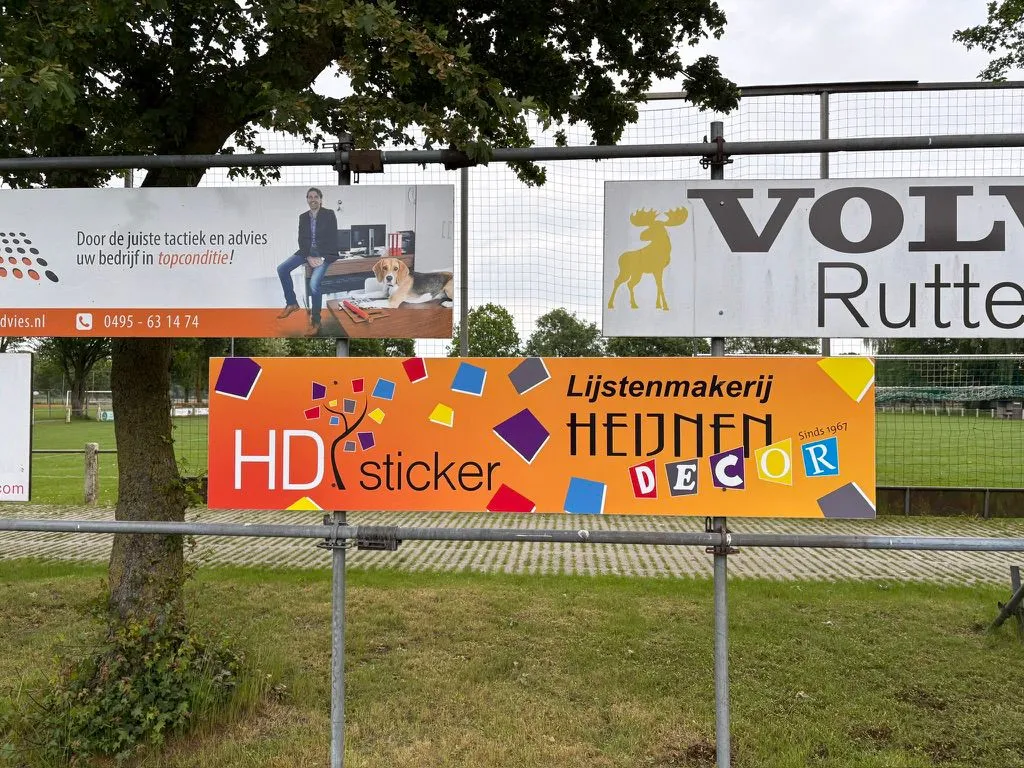 Reclameborden van sponsors aan het hek langs een voetbalveld, met het bord van HD Sticker en Heijnen Decor